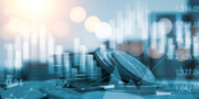 Stacks of coins are shown in the foreground with financial graphs and stock market data overlayed in the background.