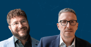 Two men in business attire pose against a blue background with faint geometric network patterns, reflecting modernes Product Management bei SupplyOn. One man is smiling with curly hair and glasses, while the other wears glasses and a neutral expression.