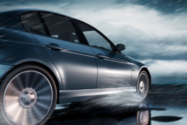 A dark-coloured car drives on a wet road, creating splashes as rain falls heavily under a cloudy sky.