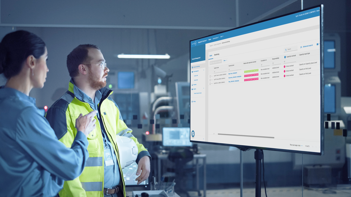 Two people in a high-tech industrial setting review data on a large monitor displaying a user interface with project status indicators.