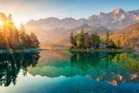 A calm mountain lake reflects pine trees and rocky peaks at sunrise, with sunlight streaming through the trees on the left.