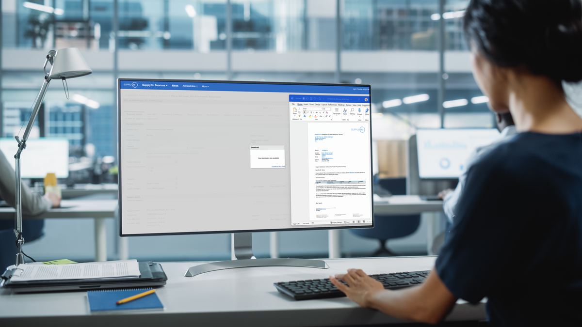 A person sits at a desk in a modern office, working on a computer with a document open on the screen. Other desks and people are visible in the background.