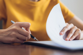 Person in a yellow shirt holds a pen and flicks through pages of a document on a table.