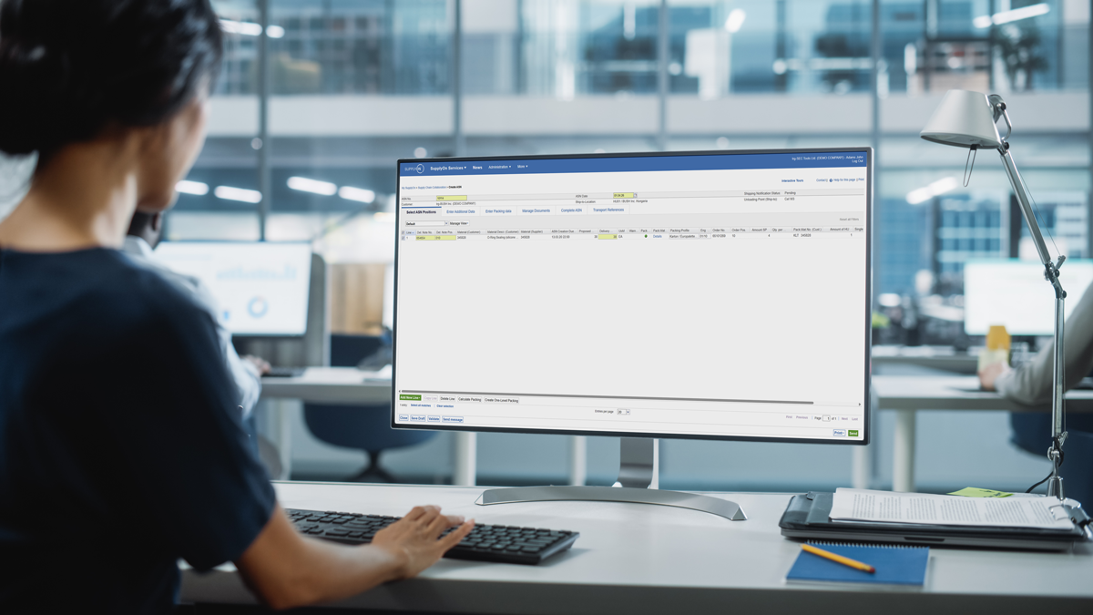 A person sits at a desk in an office, working on a desktop computer displaying a software application with tables and data fields.