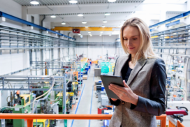 A woman in business attire stands on a balcony overlooking a large industrial factory floor, using a tablet device.