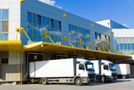 Three white lorries are parked at loading bays outside a modern commercial building with reflective glass windows and yellow awnings.