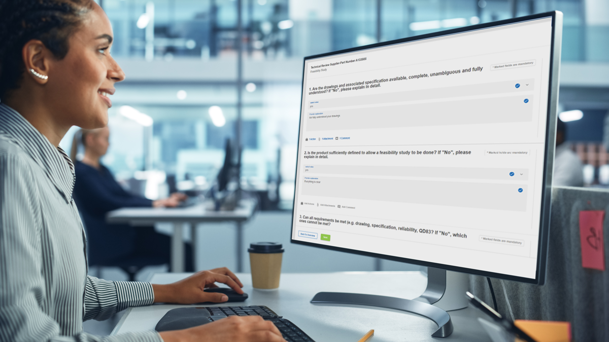 Person sits at a desk in a modern office, looking at a large computer monitor displaying a website with survey or questionnaire forms.