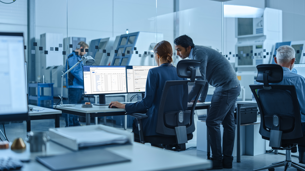 Office workers in a modern workspace look at computer monitors displaying data while others work in the background; the setting appears professional and collaborative.