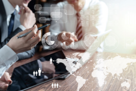 Three people in business attire sit at a table with a tablet, reviewing data visualisations and charts projected over the image, including graphs, maps, and analytics icons.