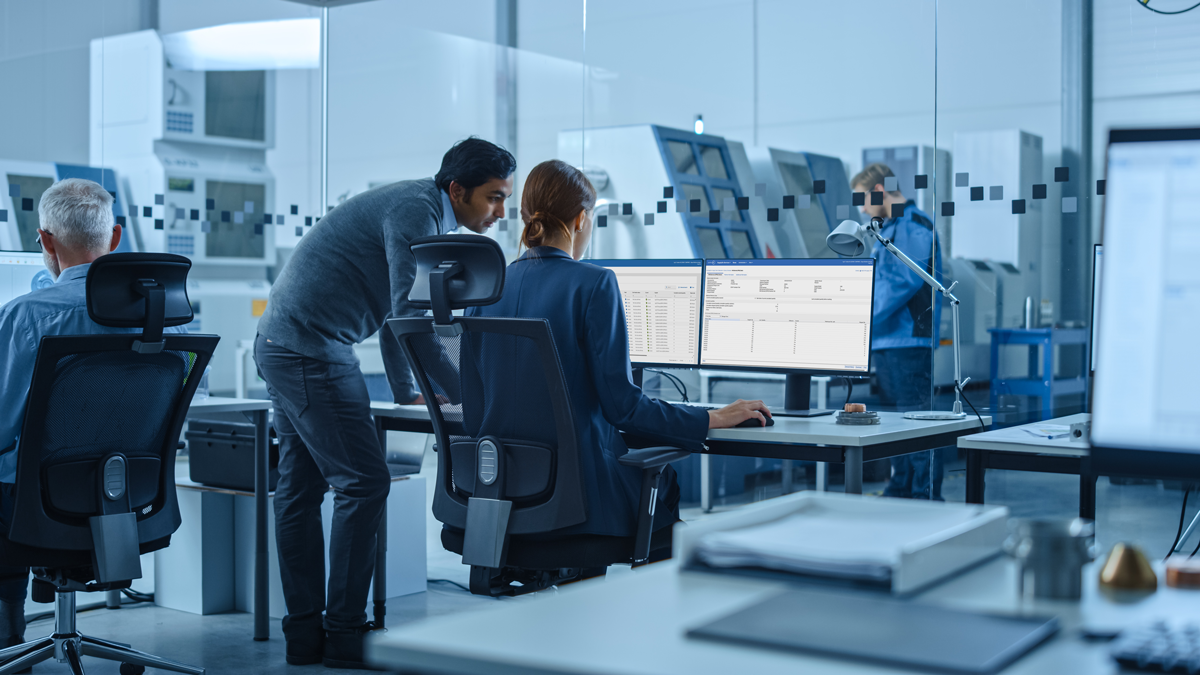 Office workers at desks monitor data on computer screens in a modern, glass-walled industrial control room; machinery and another person are visible in the background.