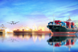 A cargo ship loaded with containers is docked at a port, with cranes, another ship, and an aeroplane flying overhead under a blue sky.