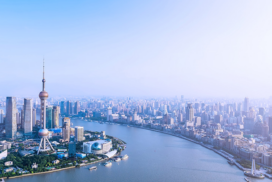 A city skyline with modern skyscrapers beside a wide river under a clear, hazy sky.