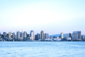 City skyline with modern high-rise buildings, viewed from across a body of water under a clear sky. Sailing boats are visible on the water.
