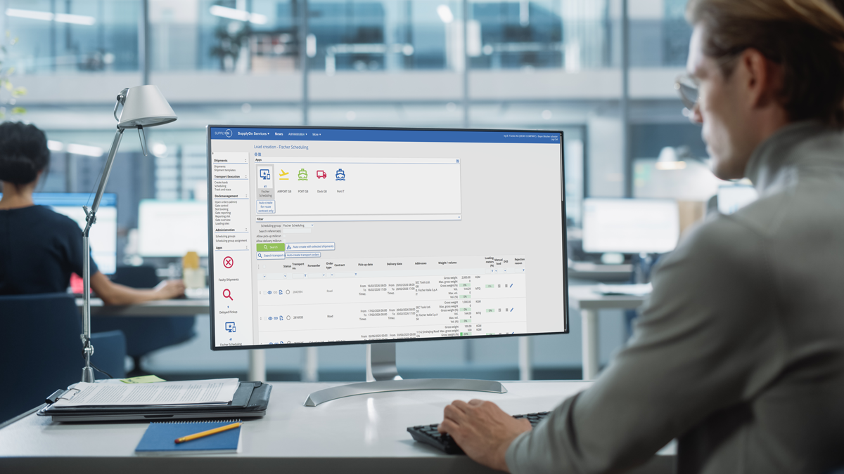 Person using a desktop computer in an office, viewing a software dashboard with task lists and icons on the screen. Other employees and desks are visible in the background.