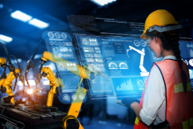 Engineer in a hard hat uses a digital touchscreen interface to monitor and control robotic arms welding in a high-tech industrial setting.