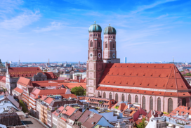 Aerial view of the Frauenkirche, a large church with twin domed towers, surrounded by red-roofed buildings in Munich, Germany, under a blue sky.