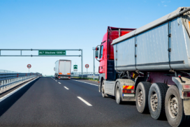 Two large lorries drive on a motorway under a clear sky, with a road sign overhead indicating ALT Stazione 1200 m.