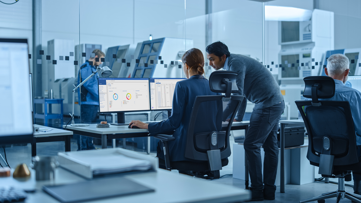 Office workers at desks view data on computer monitors, while two colleagues discuss something. Industrial equipment and another worker are visible through glass walls in the background.