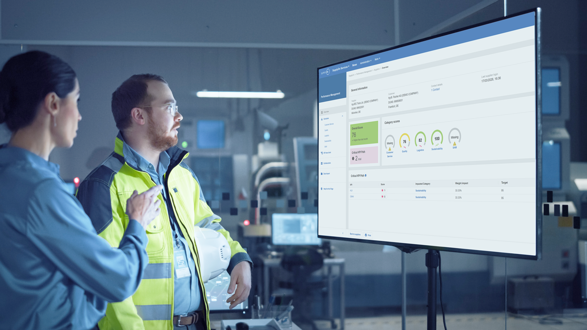 Two people in a modern industrial setting examine data and performance metrics displayed on a large monitor screen.