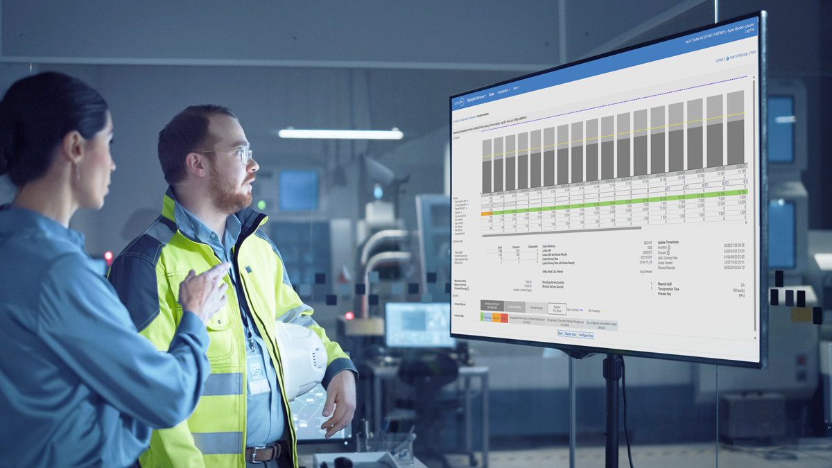 Two people in a control room analyse industrial data displayed on a large monitor, which shows a bar chart and detailed information on production performance.