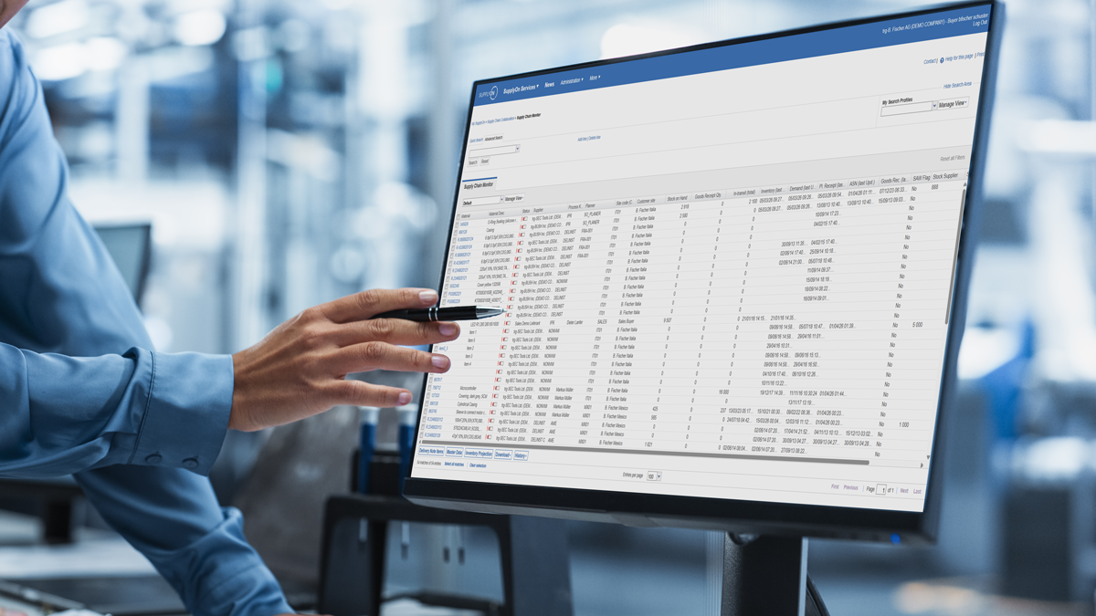 A person points a pen at a computer monitor displaying a detailed spreadsheet or data table on a business software interface in an office setting.