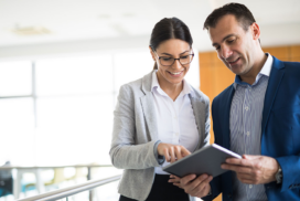 Two business professionals stand indoors, both looking at and discussing information on a tablet device.