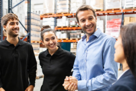 Four people stand in a warehouse, engaged in conversation and smiling. Shelves with boxes and supplies are visible in the background.