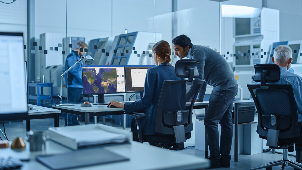 People work at computer desks in a modern office with glass walls. One screen displays a world map, and others show data. Two people are discussing something on a monitor.