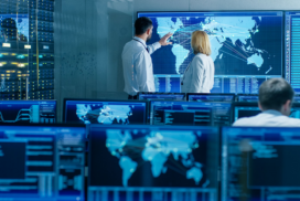Two professionals in a control room discuss data displayed on a large world map screen, surrounded by multiple computer monitors showing maps and code.