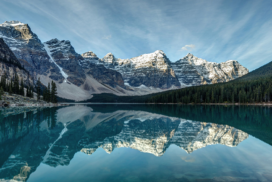 Clear lake reflecting snow-capped mountains and evergreen trees under a blue sky with wispy clouds.