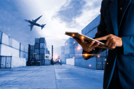 Person using a tablet in a shipping yard with cargo containers, forklift trucks, and an aeroplane flying overhead, illustrating logistics and transport.