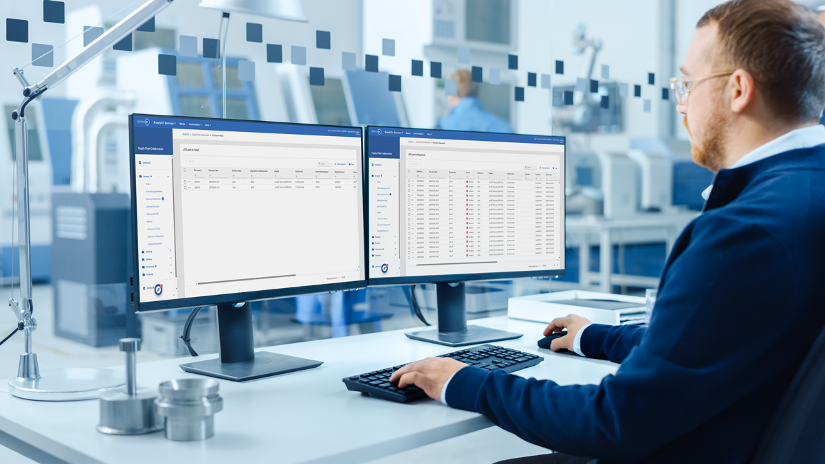 A person sits at a desk in an office, working on two computer monitors displaying data tables and graphs. Industrial equipment is visible in the background.