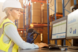 Woman in a hard hat and safety vest working at a computer workstation in a warehouse, analysing data charts on dual monitors.
