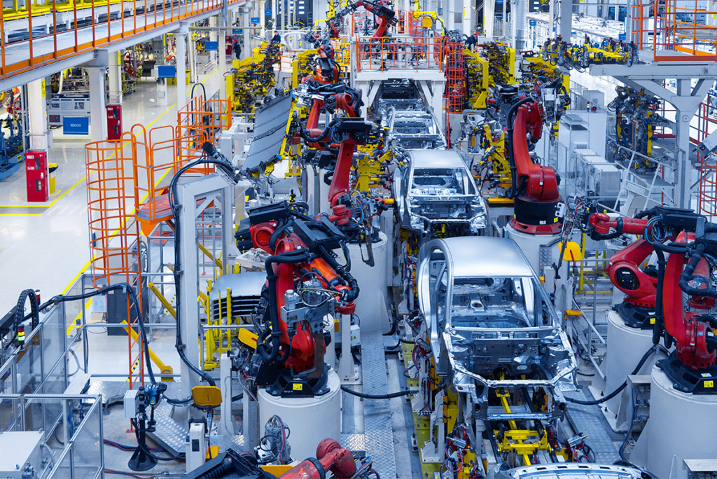 Assembly line with robotic arms assembling car bodies in a modern, brightly lit automotive manufacturing facility.