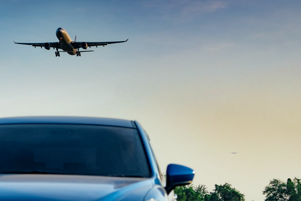 A commercial aeroplane approaches for landing in the sky above a blue car parked near trees at sunset.