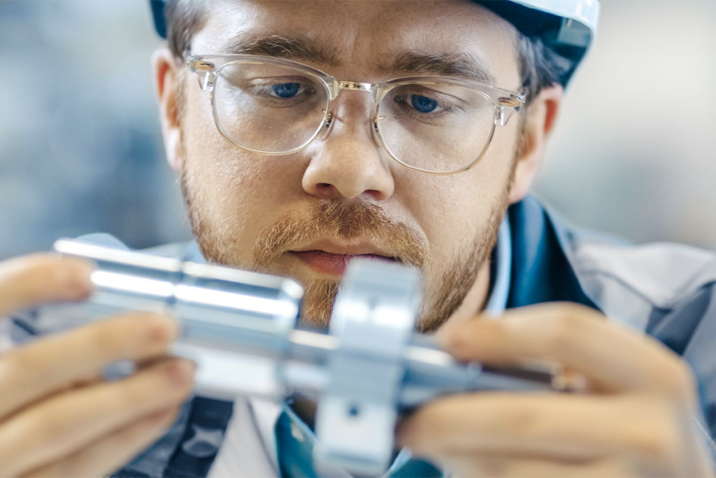 A man wearing glasses and a hard hat closely examines a metal cylindrical component, likely in an industrial or manufacturing setting.
