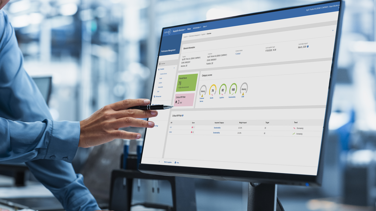 Person pointing a pen at a computer monitor displaying a dashboard interface with charts, status indicators, and data tables in an office or industrial setting.
