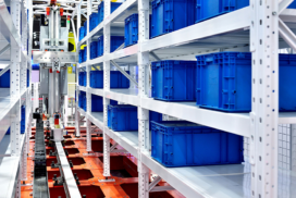 Metal shelves filled with blue plastic bins in a warehouse; an automated robotic system operates between the shelves.