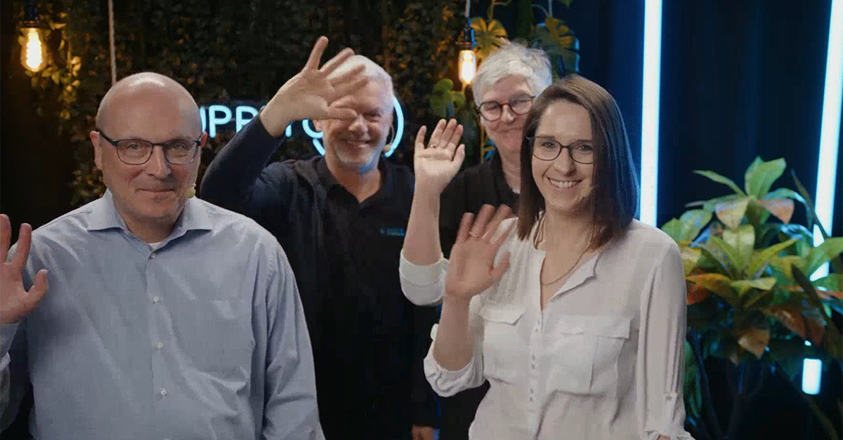 Four adults, three men and one woman, stand indoors and wave at the camera, smiling. There are plants and illuminated vertical lights in the background, reflecting a friendly team spirit within the supplier network.
