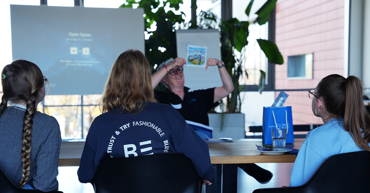 A person gives a presentation to three seated attendees, holding up a booklet, with a projection screen and a large plant in the background.