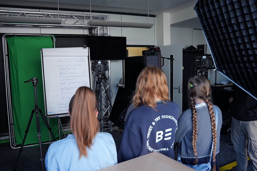 Three people with long hair face a whiteboard with handwritten notes in a studio setup, surrounded by cameras, lights, and a green screen—capturing all the creative energy of a girls' day on set.