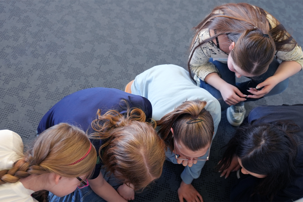 Five girls with long hair, seen from above, are sitting close together on a patterned carpet during a girls’ day, looking down at something out of view.