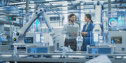 Two engineers discuss data in a high-tech factory, surrounded by robotic arms, conveyors, and digital screens displaying charts and graphs.