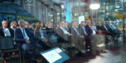 A group of people in business attire are seated in rows at a conference or seminar in a modern, glass-walled venue.
